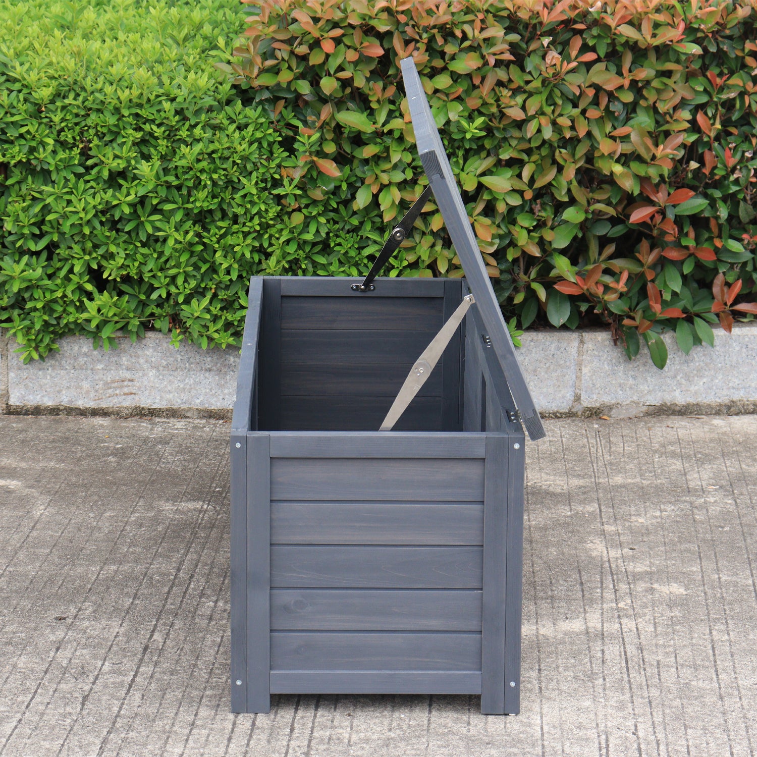 A charcoal wooden storage box with its lid propped open using two metal support rods, set outdoors on a concrete surface with green bushes in the background.