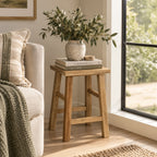 A small wooden stool with a rustic finish stands beside a light beige sofa in a bright room. On top of the stool are two stacked books and a textured ceramic vase filled with leafy green stems.