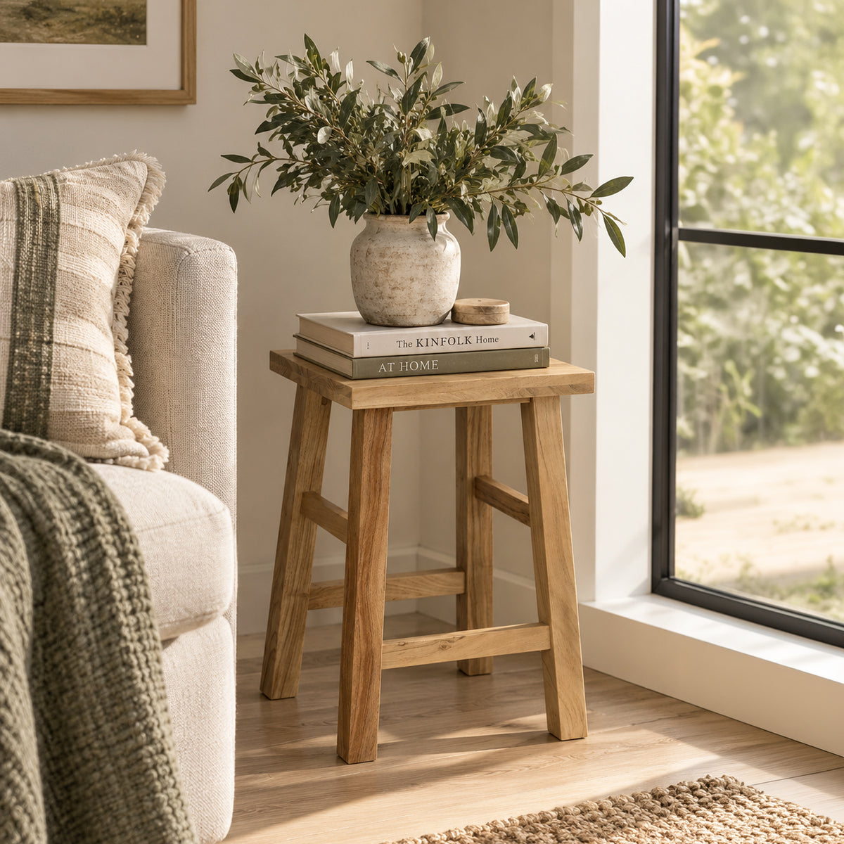 A small wooden stool with a rustic finish stands beside a light beige sofa in a bright room. On top of the stool are two stacked books and a textured ceramic vase filled with leafy green stems.