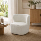 A modern barrel-style accent chair upholstered in a chenille fabric in cream, placed in a warm, minimalist living room setting with a wooden sideboard, beige rug, and soft natural light.