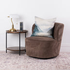 A brown textured swivel armchair with a blue and white mountain landscape cushion, styled next to a small wooden side table with a brass lamp in a bright room.
