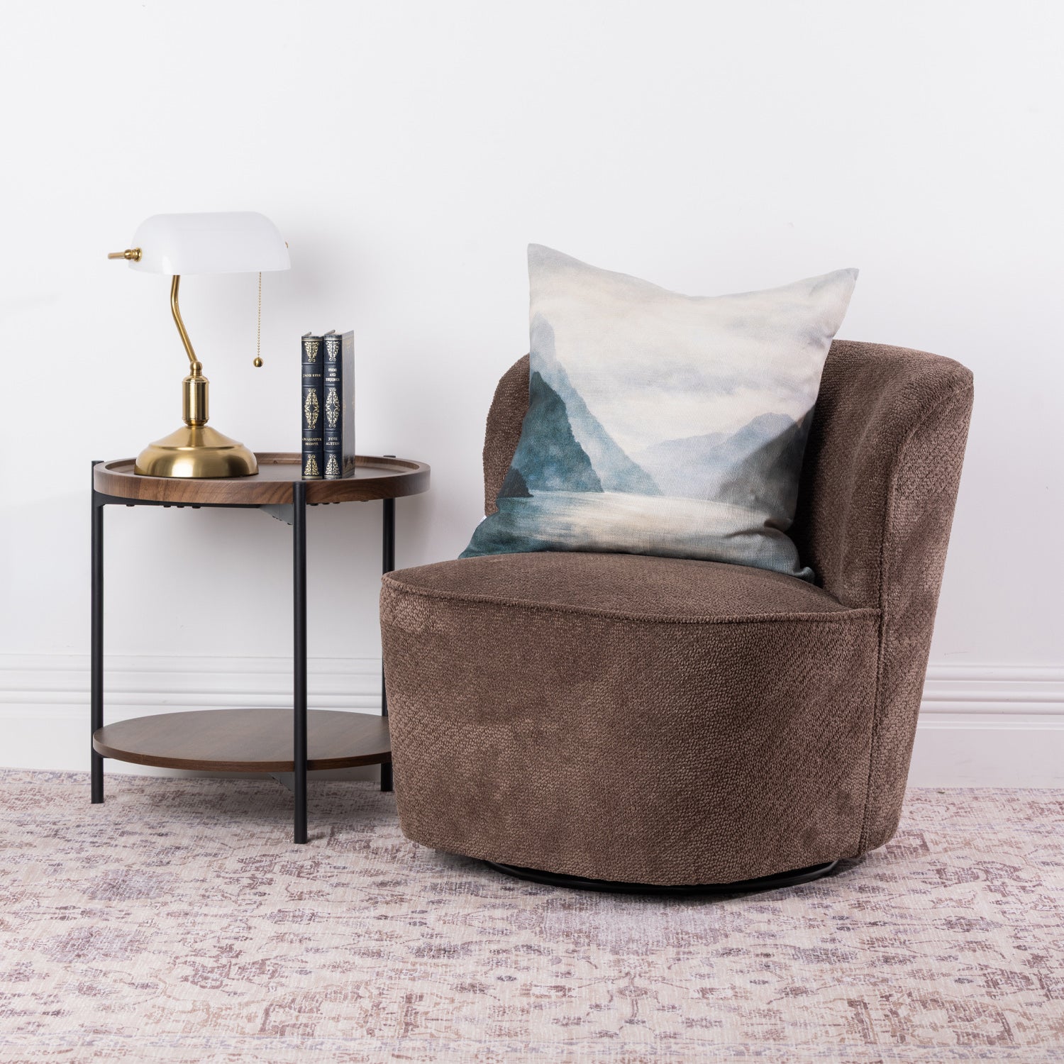 A brown textured swivel armchair with a blue and white mountain landscape cushion, styled next to a small wooden side table with a brass lamp in a bright room.