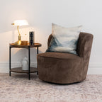 A brown upholstered swivel armchair with a blue-gray mountain landscape cushion, styled next to a small wooden and metal side table with a lamp and books in a bright room.