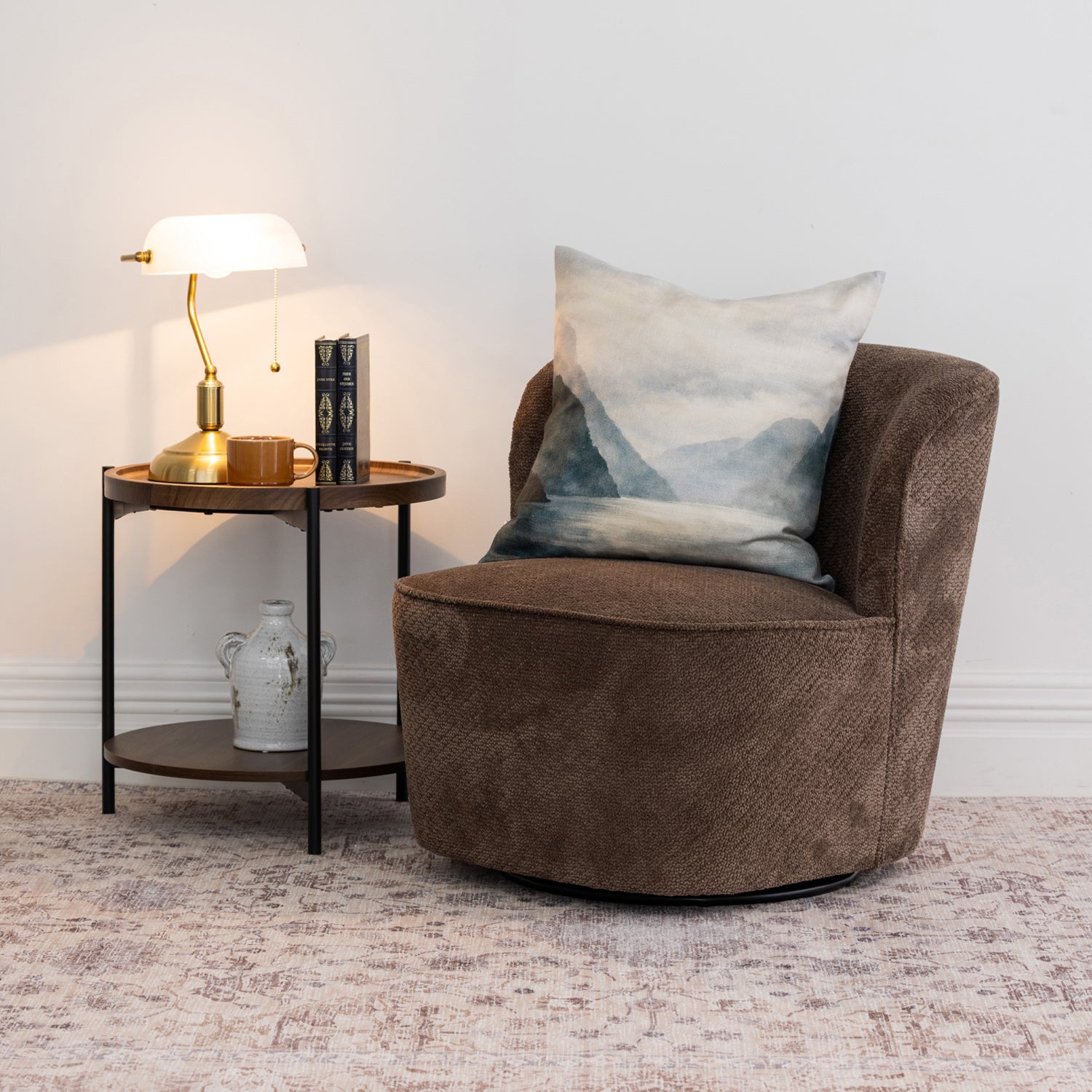 A brown upholstered swivel armchair with a blue-gray mountain landscape cushion, styled next to a small wooden and metal side table with a lamp and books in a bright room.