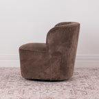 A brown chenille textured fabric swivel armchair with a rounded backrest, shown on a distressed patterned rug against a white wall.