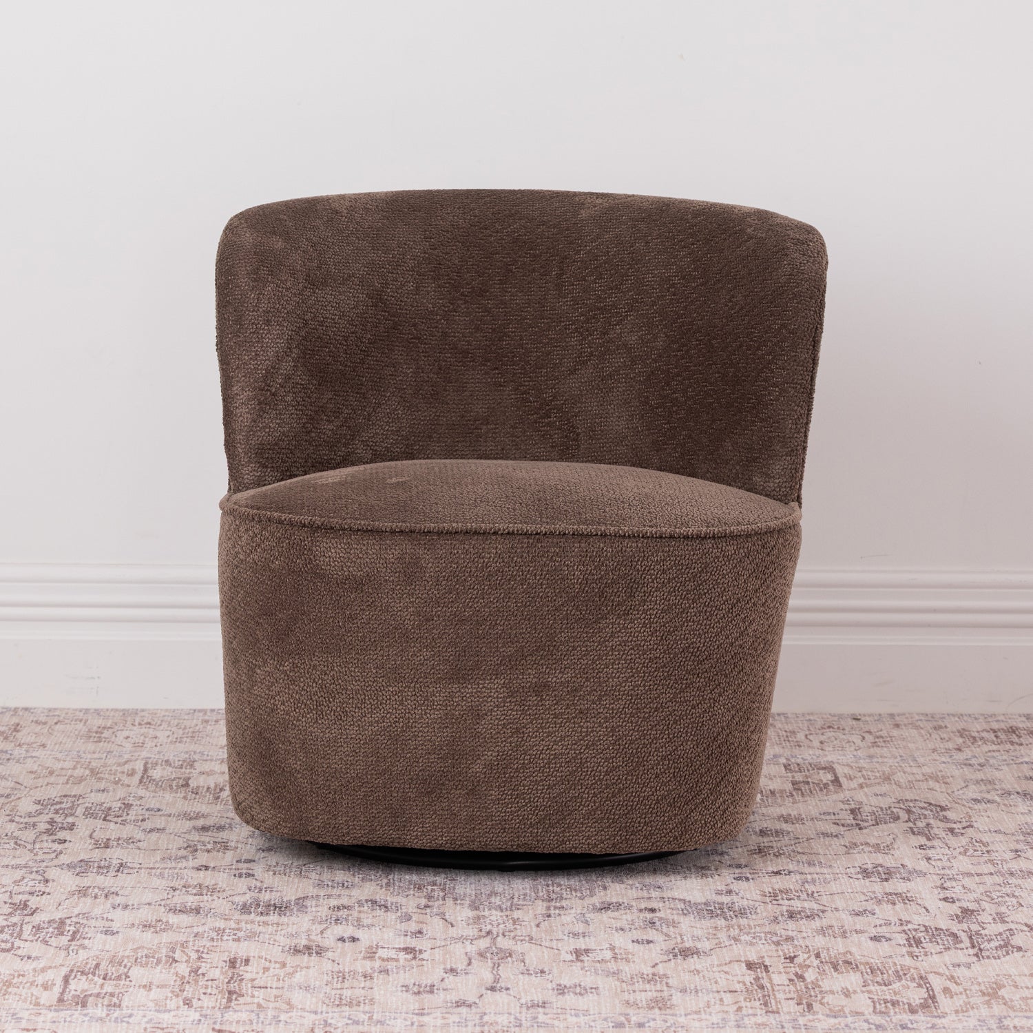 A brown chenille textured fabric swivel armchair with a rounded backrest, shown on a distressed patterned rug against a white wall.