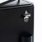 Detailed close-up of the silver metal bottle opener mounted on the side of the matte black cooler box, with the built-in bottle cap catcher positioned below.