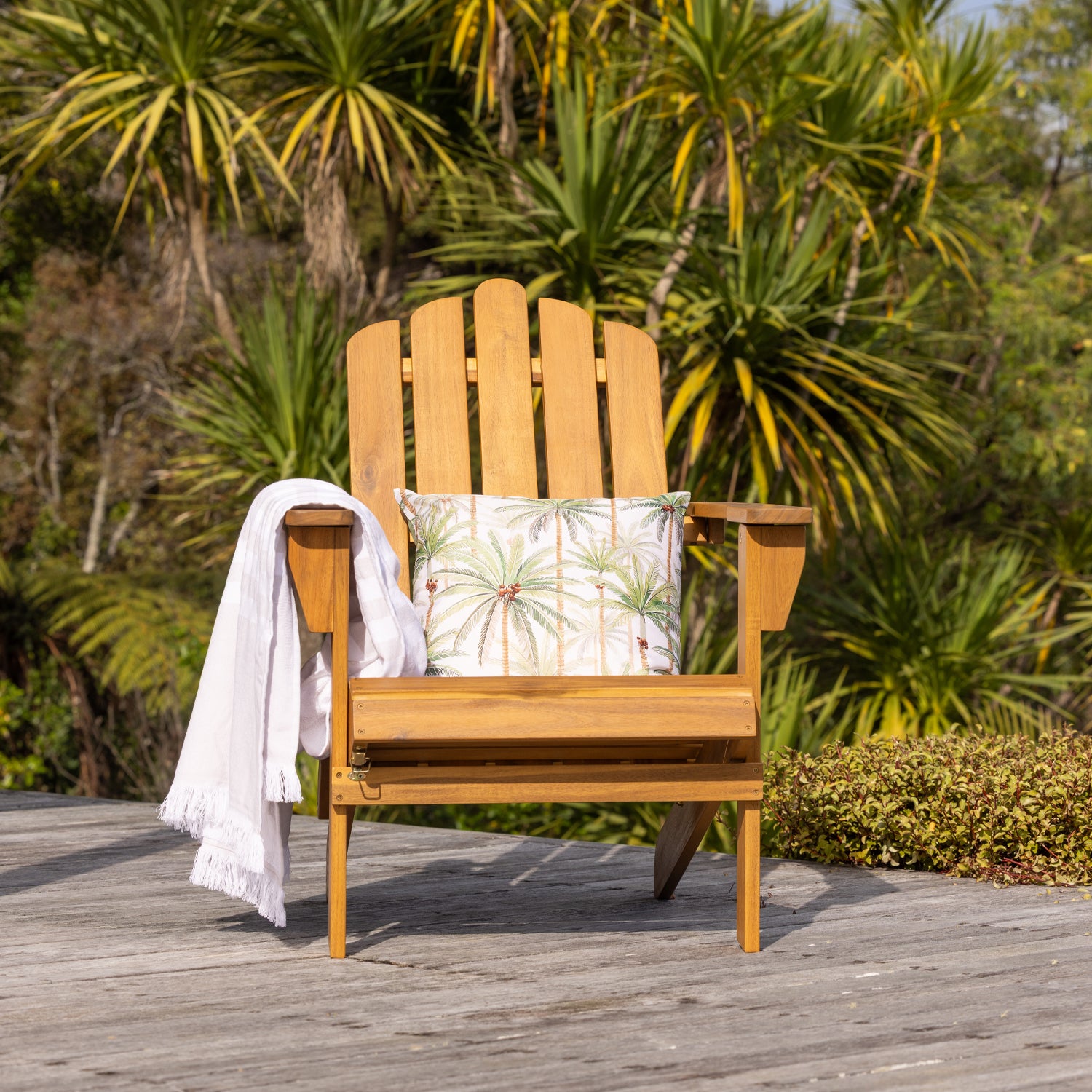A wooden cape cod-style outdoor chair sits on a weathered deck with tropical palm foliage in the background.