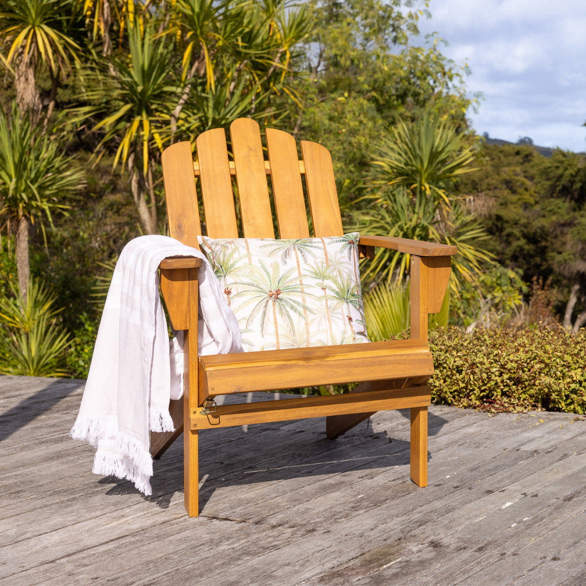 A wooden cape cod-style outdoor chair sits on a weathered deck with tropical palm foliage in the background.