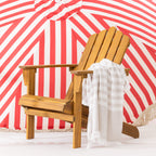 A wooden cape cod chair with a striped fringed towel draped over the arm, set against a red and white striped umbrella backdrop.
