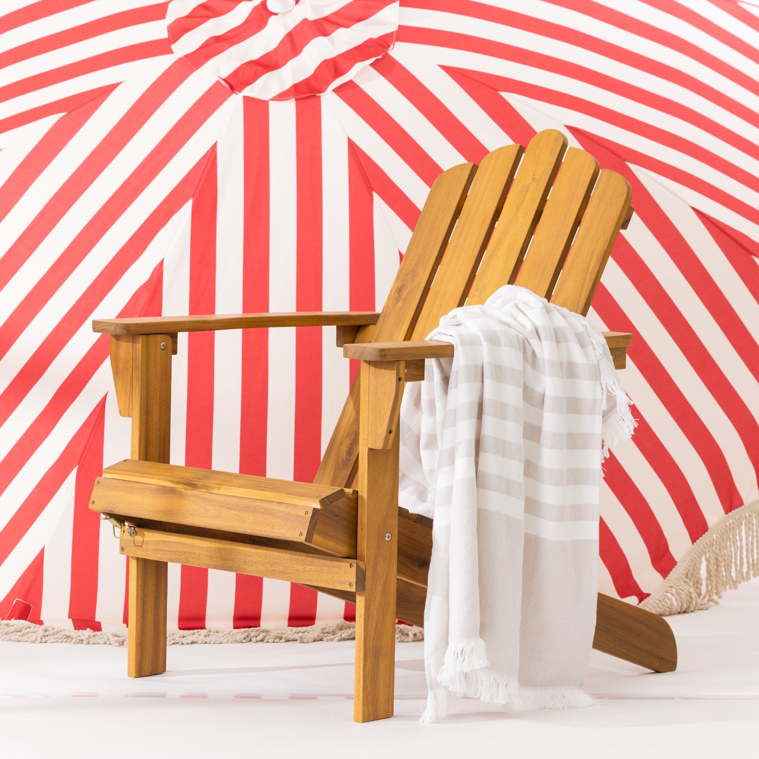 A wooden cape cod chair with a striped fringed towel draped over the arm, set against a red and white striped umbrella backdrop.