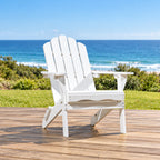A white outdoor Adirondack chair sits on a wooden deck with a grassy lawn and ocean view in the background.
