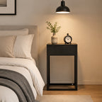 A modern black nightstand in a bedroom setting, styled with a small vase of greenery and a black alarm clock, next to a bed with white linens under a black pendant lamp.