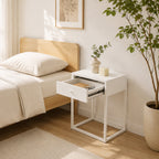 A modern white nightstand with an open drawer and square metal legs, styled next to a bed in a bright, minimalist bedroom with a potted plant.