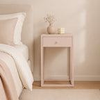 A light blush pink nightstand with a single drawer and open lower shelf, styled in a bedroom next to a bed with neutral bedding. Decorative items on top include a small vase with dried stems and a textured cream candle.
