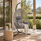 A woven hanging egg chair with gray cushions and a cream throw blanket sits on a patio porch with a wooden post, plants, and outdoor greenery in the background.