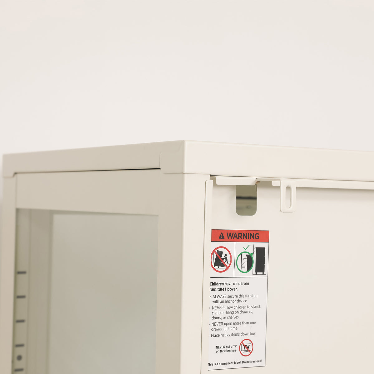 Close-up of the white shelving cabinet's rear corner, showing the furniture anti-tip bracket and safety warning label.