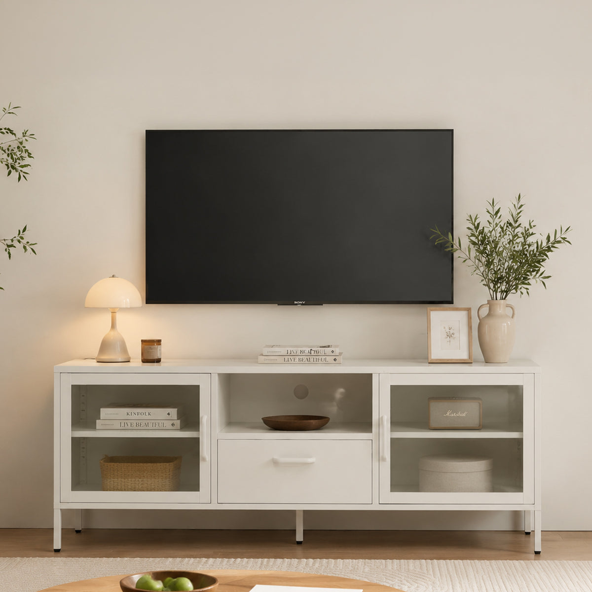 White TV cabinet styled in a modern living room beneath a wall-mounted television, featuring glass doors, central drawer, open media shelf and decorative accessories.