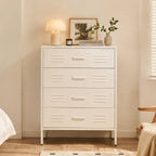 A white four-drawer chest of drawers with decorative horizontal vent-like cutouts on each drawer front, shown in a softly styled bedroom setting with a lamp, framed art, books, and small plants on top.