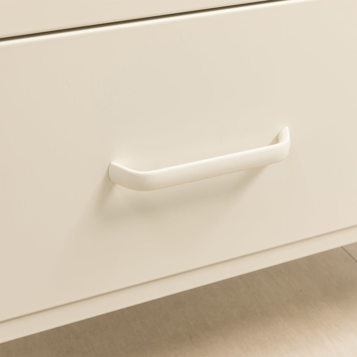 A close-up of a minimalist, curved drawer pull handle in a warm off-white finish mounted on a matching cream-colored drawer front.
