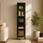 A tall black narrow cabinet with glass doors stands in a styled living room corner. The cabinet has four open shelves and a lower woven storage basket, styled with books, vases, a bust, and decor.