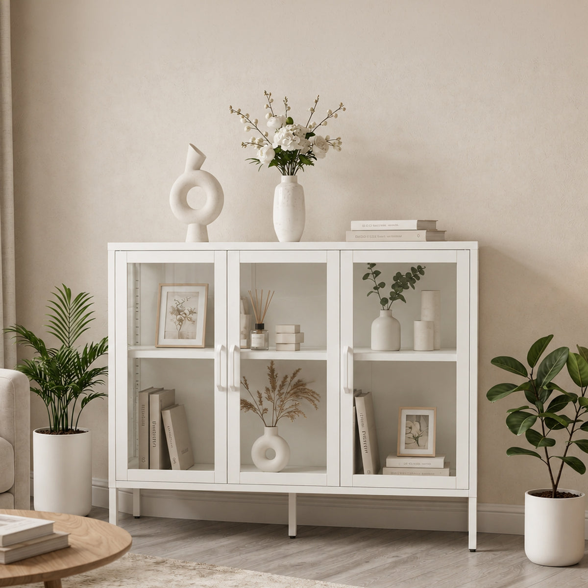 A white three-door glass-front sideboard cabinet styled in a minimalist living room setting with books, vases, and potted plants.