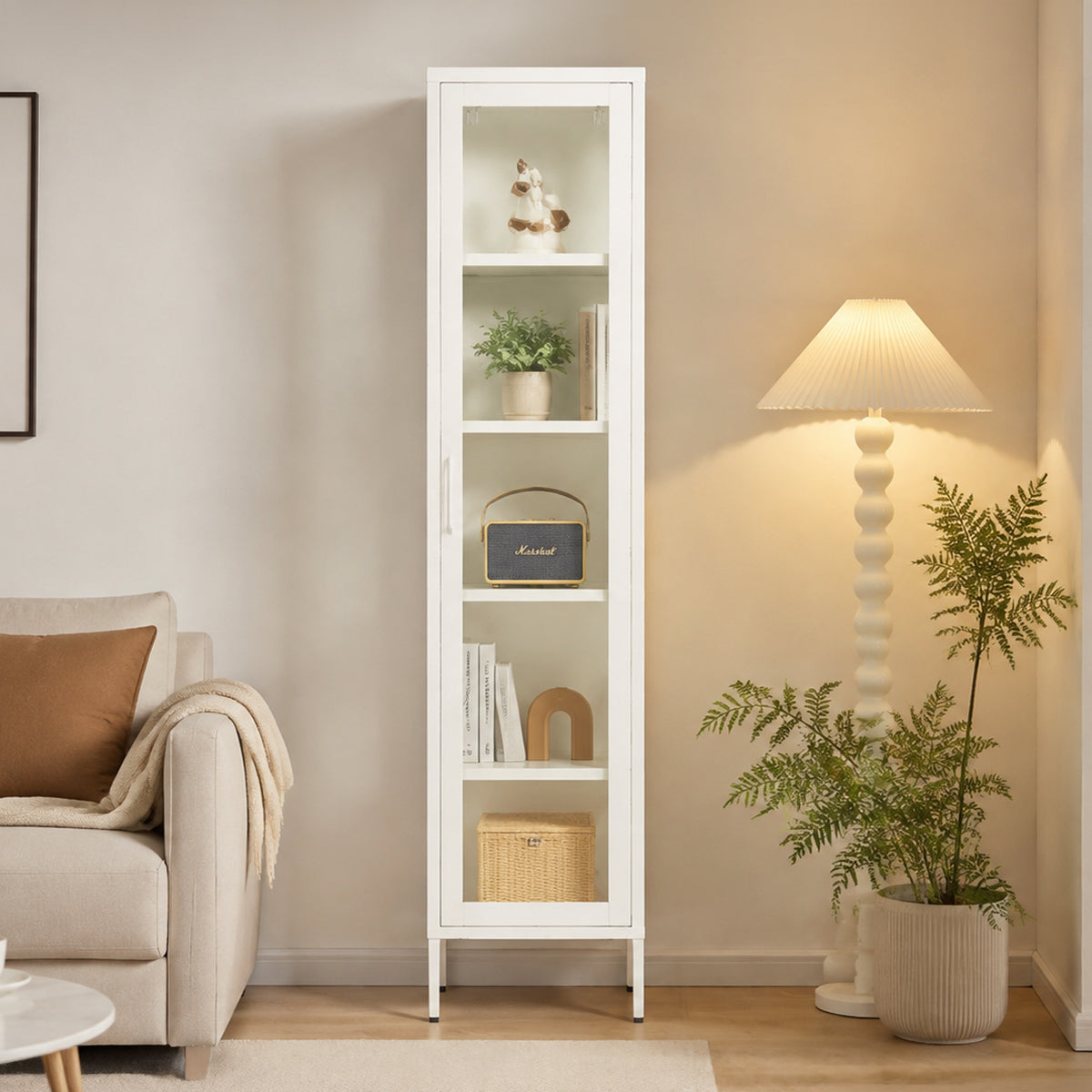 A tall white narrow shelving cabinet with four glass-front shelves stands in a styled living room next to a beige armchair, a floor lamp, and potted plants.