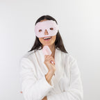 A woman wearing a white textured robe and a light pink eye mask holds a matching pink skincare sponge against a plain white background.