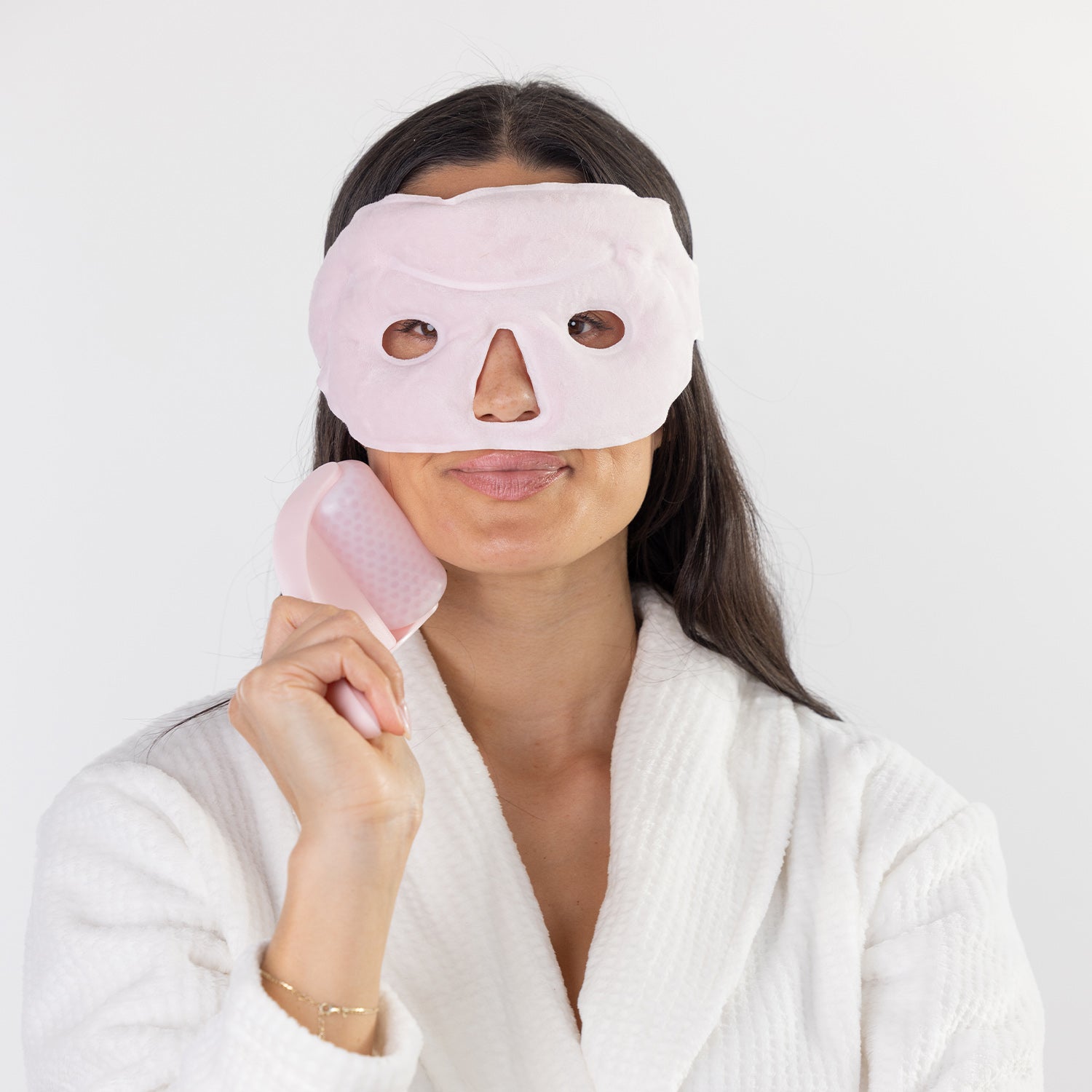 A woman wearing a white textured robe uses a light pink handheld facial massage device on her face, with a matching pink sheet mask applied. Shot against a clean white background.