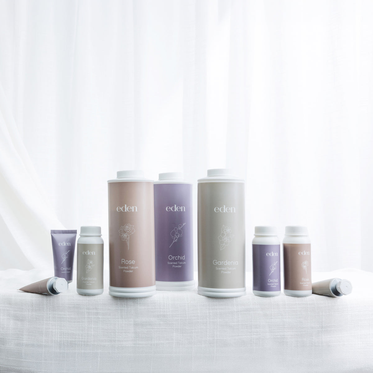 Eden Rose, Gardenia and Orchid scented talcum powder and hand cream range displayed together against a soft white curtain backdrop.