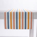 A woven table runner with vertical stripes in blue, orange, and dusty pink on an off-white background, draped over a white wooden table.