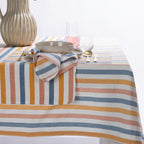 A striped tablecloth with horizontal bands of blue, mustard yellow, peach, and cream, styled on a table with a matching napkin, plates, cutlery, and glassware.