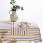 A striped tablecloth in pastel blue, orange, yellow, and white, styled on a dining table with a matching striped napkin, a ceramic bowl, gold-toned cutlery, and a ribbed beige pitcher vase with green foliage.