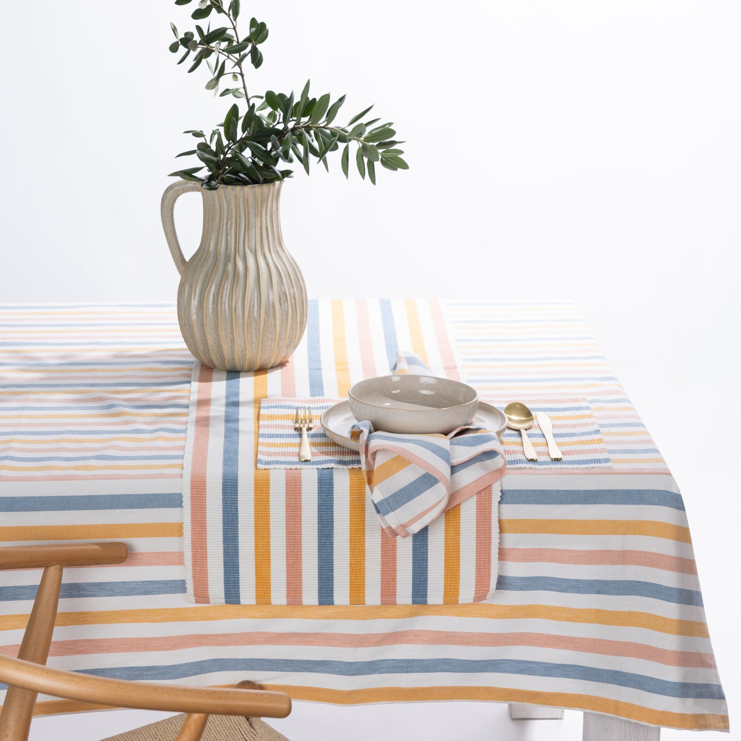 A striped tablecloth in pastel blue, orange, yellow, and white, styled on a dining table with a matching striped napkin, a ceramic bowl, gold-toned cutlery, and a ribbed beige pitcher vase with green foliage.