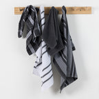 Home Co. Terry Tea Towels 4 Pack in Charcoal, shown hanging on a wooden wall rack with coordinating check, stripe, and plain charcoal designs.