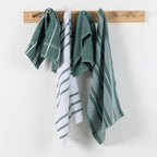 Home Co. Terry Tea Towels 4 Pack in Sage, shown hanging on a wooden wall rack with coordinating check, stripe, and plain sage designs.