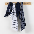 Home Co. Terry Tea Towels 4 Pack in Navy, shown hanging on a wooden wall rack with coordinating check, stripe, and plain navy designs.