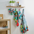 Design Plus Kiwiana Pohutukawa kitchen textile collection displayed hanging on wall hooks beneath a wooden shelf, including an apron, oven mitt, double oven glove and tea towel, showcasing vibrant pōhutukawa floral prints in a contemporary kitchen setting.