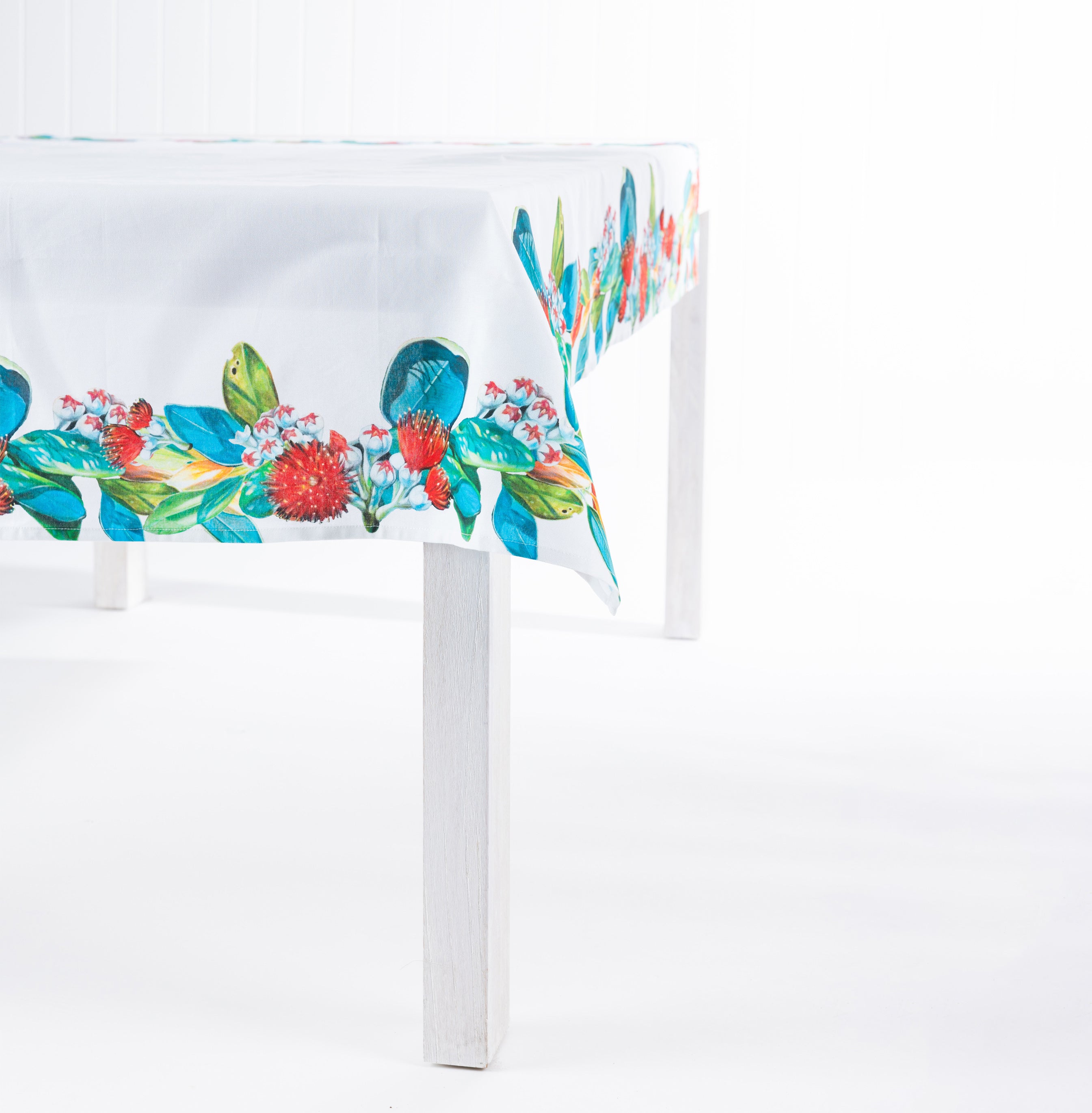 Design Plus Kiwiana Pohutukawa Tablecloth featuring a vibrant pōhutukawa floral border with rich green foliage on a crisp white tablecloth, draped over a white dining table in a clean studio setting.