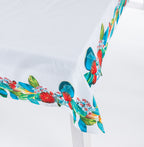 Close-up of the Design Plus Kiwiana Pohutukawa Tablecloth draped over the corner of a white dining table, highlighting the vibrant pōhutukawa floral border with lush green leaves and crisp white centre.