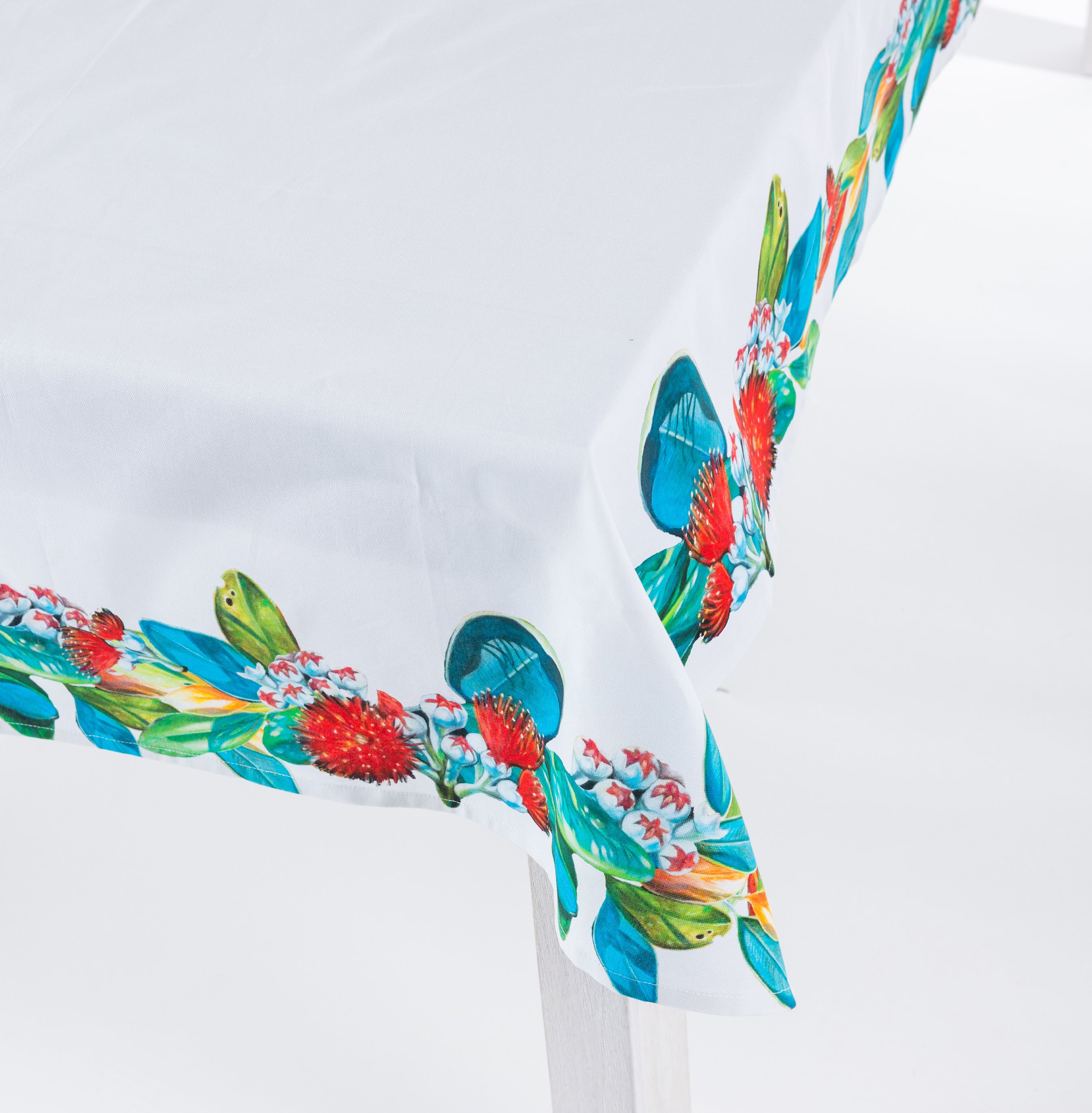 Close-up of the Design Plus Kiwiana Pohutukawa Tablecloth draped over the corner of a white dining table, highlighting the vibrant pōhutukawa floral border with lush green leaves and crisp white centre.