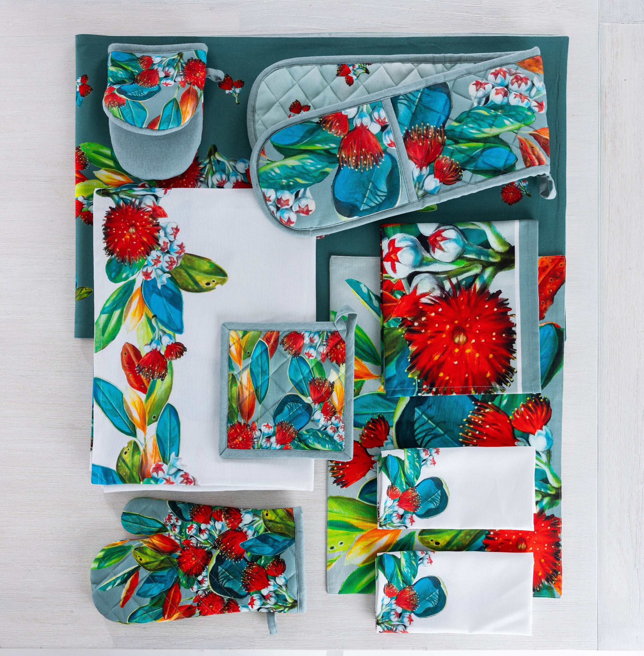 Design Plus Kiwiana Pohutukawa kitchen textile collection displayed flat, including an apron, oven mitt, double oven glove, pot holder, tea towel, table runner and napkins, all featuring colourful pōhutukawa floral designs in coordinating sage, white and vibrant botanical prints.