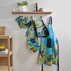 Design Plus Kiwiana Kowhai kitchen textile collection displayed hanging on wall hooks beneath a wooden shelf, including a full apron, half apron, oven mitt, double oven glove and tea towel, all featuring colourful tūī bird and kōwhai flower prints in a contemporary kitchen setting.