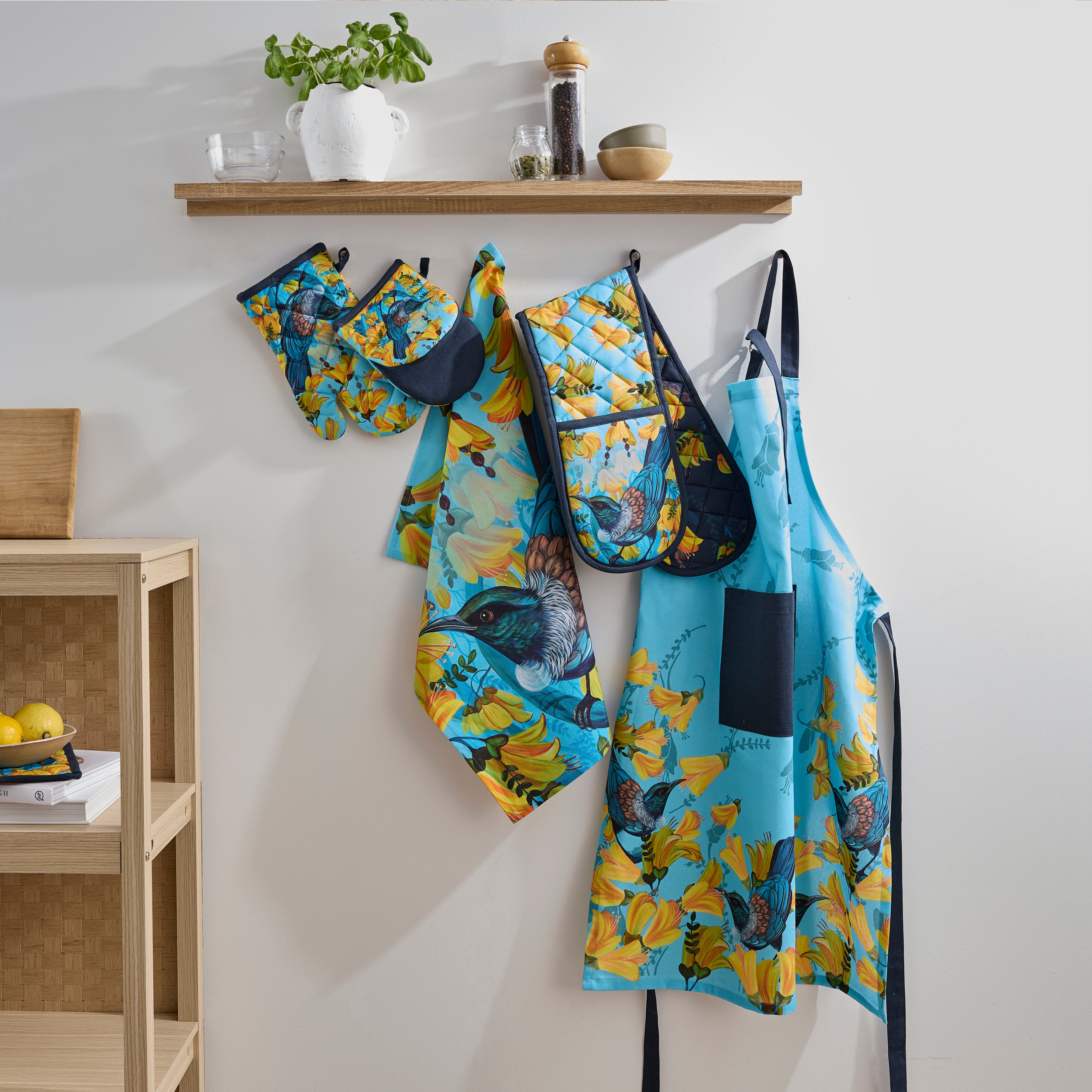 Design Plus Kiwiana Kowhai kitchen textile collection displayed hanging on wall hooks beneath a wooden shelf, including a full apron, half apron, oven mitt, double oven glove and tea towel, all featuring colourful tūī bird and kōwhai flower prints in a contemporary kitchen setting.