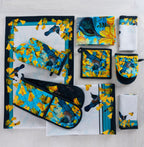 Design Plus Kowhai kitchen textile collection displayed flat, including a table runner, oven mitt, double oven glove, pot holder, tea towels and napkins, all featuring colourful tūī birds and yellow kōwhai flowers on coordinating blue and white designs.