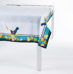 Design Plus Kowhai Tablecloth in Yellow featuring vibrant kōwhai blossoms and tūī birds along a decorative border, draped over a white dining table with a soft blue centre and New Zealand-inspired botanical design.