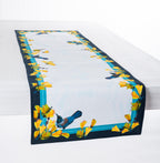 Design Plus Kowhai Table Runner featuring a border of vibrant yellow kōwhai flowers and blue tūī birds on a soft white background, displayed draped along a white timber table.