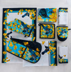 Design Plus Kowhai kitchen textile collection displayed flat, including a table runner, oven mitt, double oven glove, pot holder, tea towels and napkins, all featuring colourful tūī birds and yellow kōwhai flowers on coordinating blue and white designs.