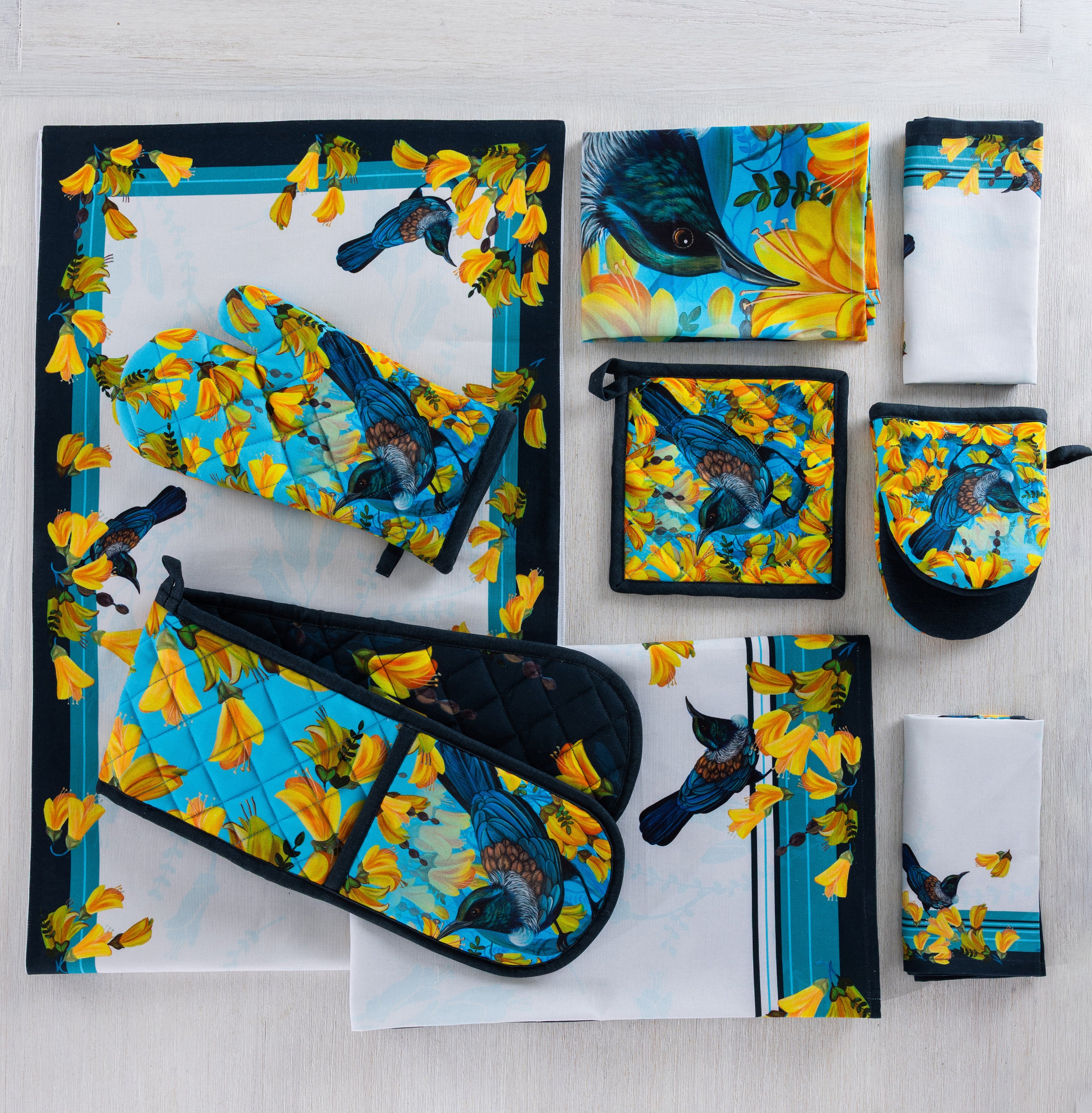 Design Plus Kowhai kitchen textile collection displayed flat, including a table runner, oven mitt, double oven glove, pot holder, tea towels and napkins, all featuring colourful tūī birds and yellow kōwhai flowers on coordinating blue and white designs.