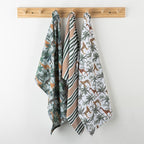 Three patterned kitchen towels hanging from a wooden peg rack on a white wall. The towel on the left has a green jungle print with orange animals, the middle towel has green and white diagonal stripes with orange stripes, and the towel on the right has a white background with green palm trees and orange animals.